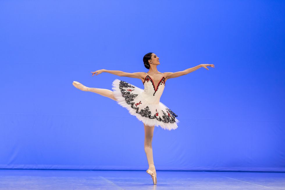 Carolina Pires does a piqu\u00e9 first arabesque on pointe on her left leg. She smiles brightly, wearing a white tutu with black and red trim, pink tights and pointe shoes. Her dark brown hair is pulled into a low bun.