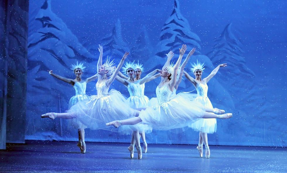 Two ballerinas in long white tutus and spkiy white crowns leap through the air with thier arms in a V-shape. Behind them, four similarly dressed dancers bour\u00e9e in sous-sus.
