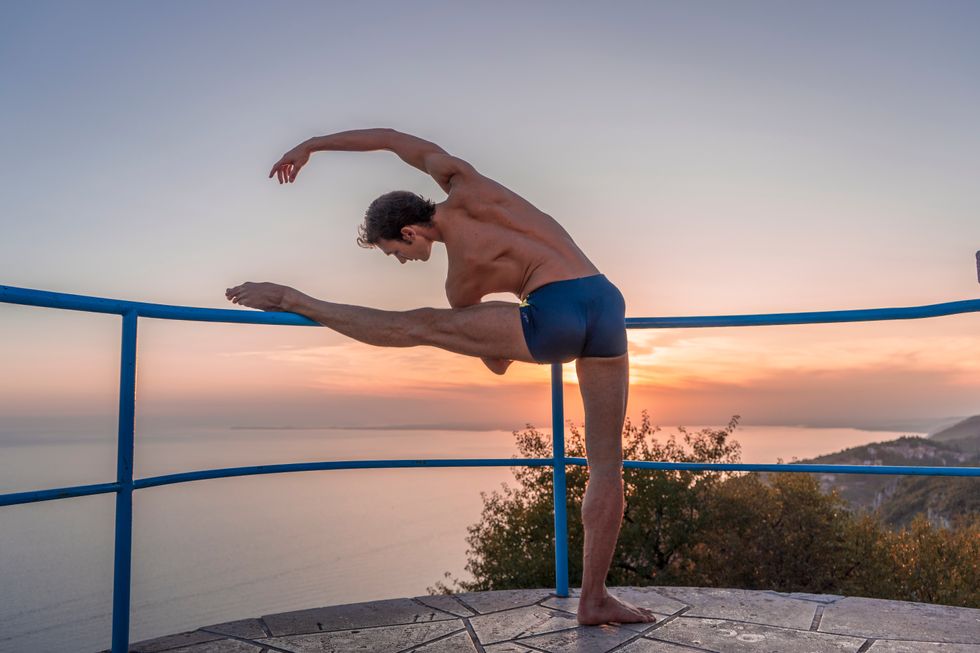 A young male ballet dancer in blue shorts stretches with his left leg on an outdoor bannister overlooking the sea.