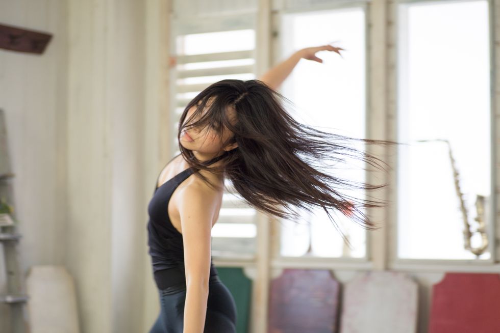 An Asian female dancer in a black tank top and leggings is shown from the thigh up in an apartment, dancing freely with her hair down and her right arm reaching high to the side of her.
