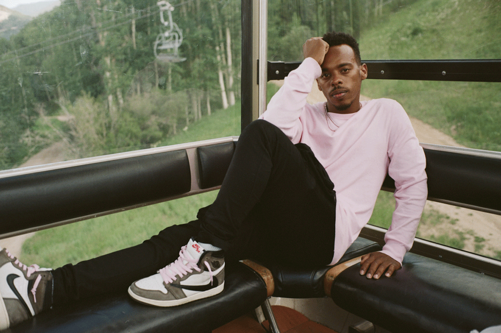 Lil Buck, in a pink sweatshirt, black pants, and white and gray sneakers with pink laces, casually reclines inside a glass-enclosed ski gondola as it heads up a mountain