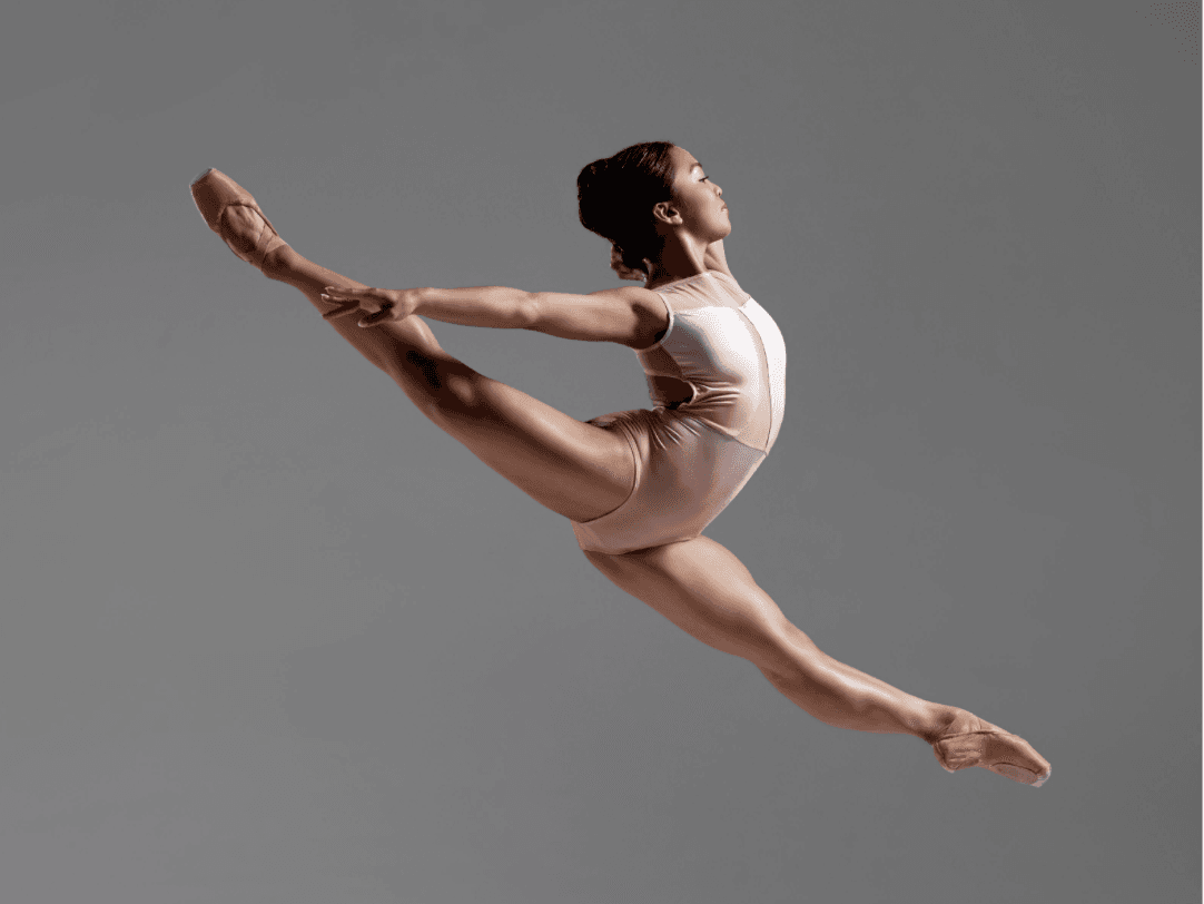 A dancer in a light leotard jumps in a wide split leap, her back leg reaching up on a diagonal, one arm behind her head. 