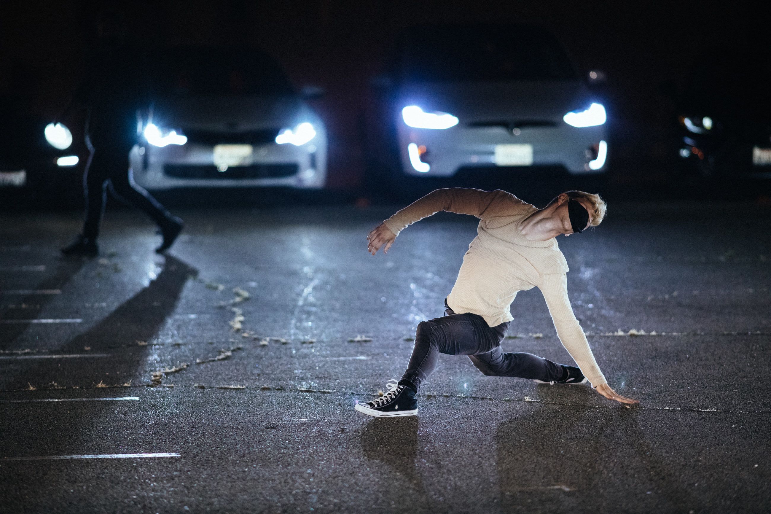 A male dancer in jeans and sneakers crouches in a wide lunge, one hand reaching back. He's wearing a mask, and car headlights illuminate him.