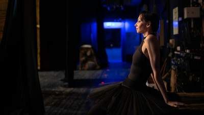 A ballerina sits backstage in a black tutu looking at the stage from the wings.