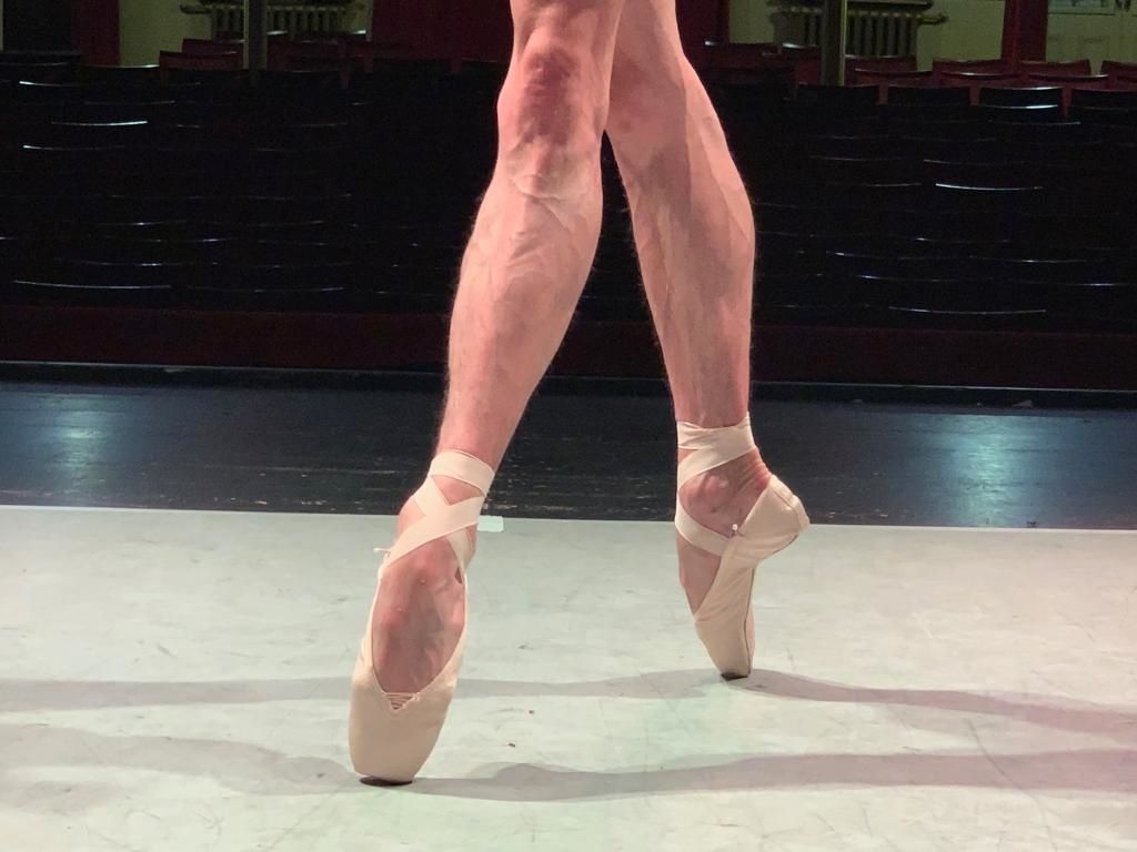 A close up of a man's lower legs and feet standing in fourth position on pointe