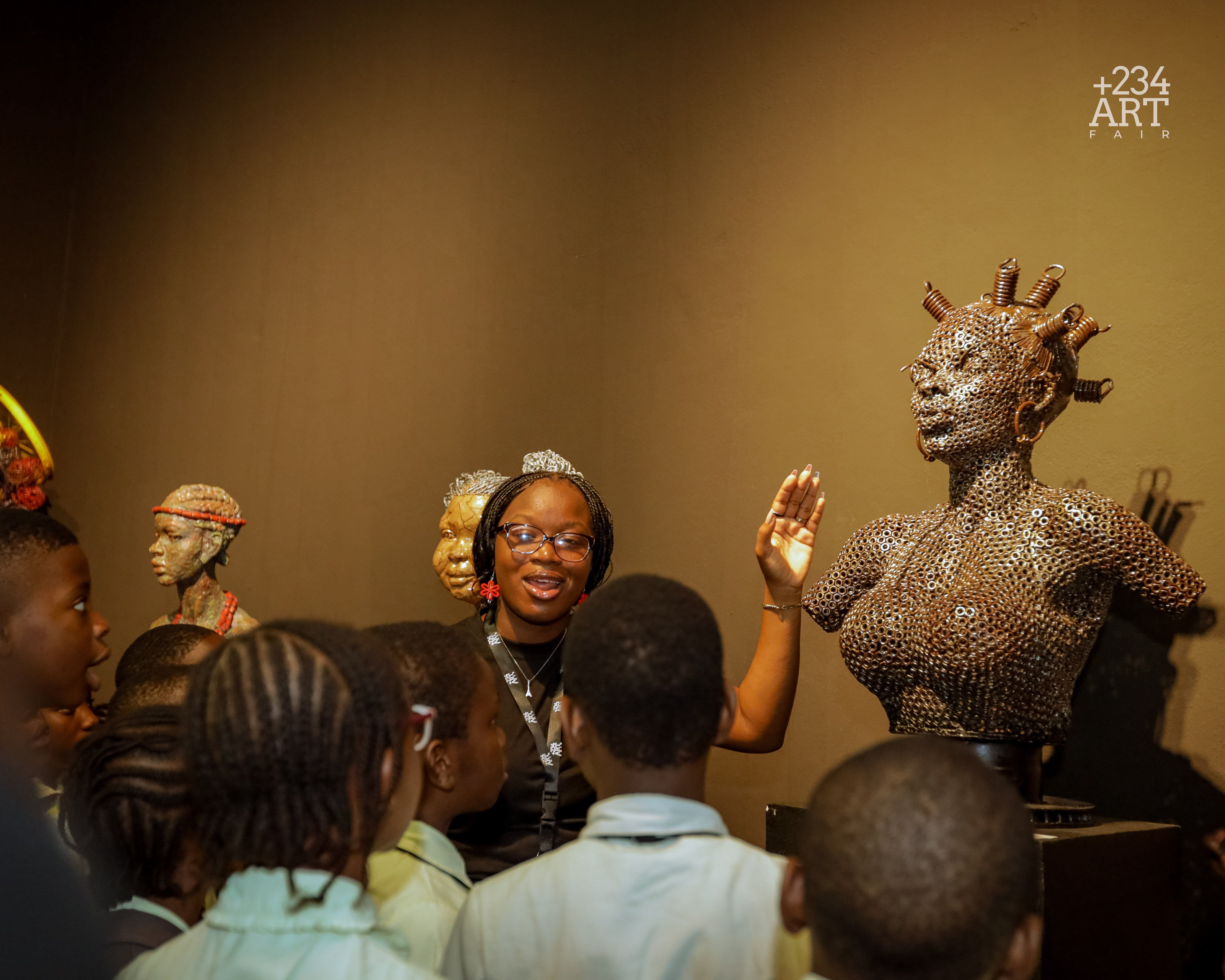 A smiling woman stands in front of a group of schoolchildren, explaining a striking female bust sculpture made from metal washers and coils at an art fair, with other colorful, textured sculptures displayed behind her.