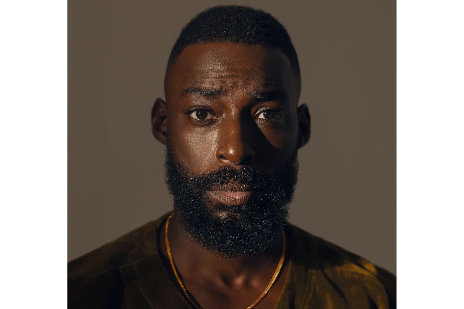 Chef Eric Adjepong looking straight into the camera. He wears a brown shirt and a gold chain. He has a full beard.