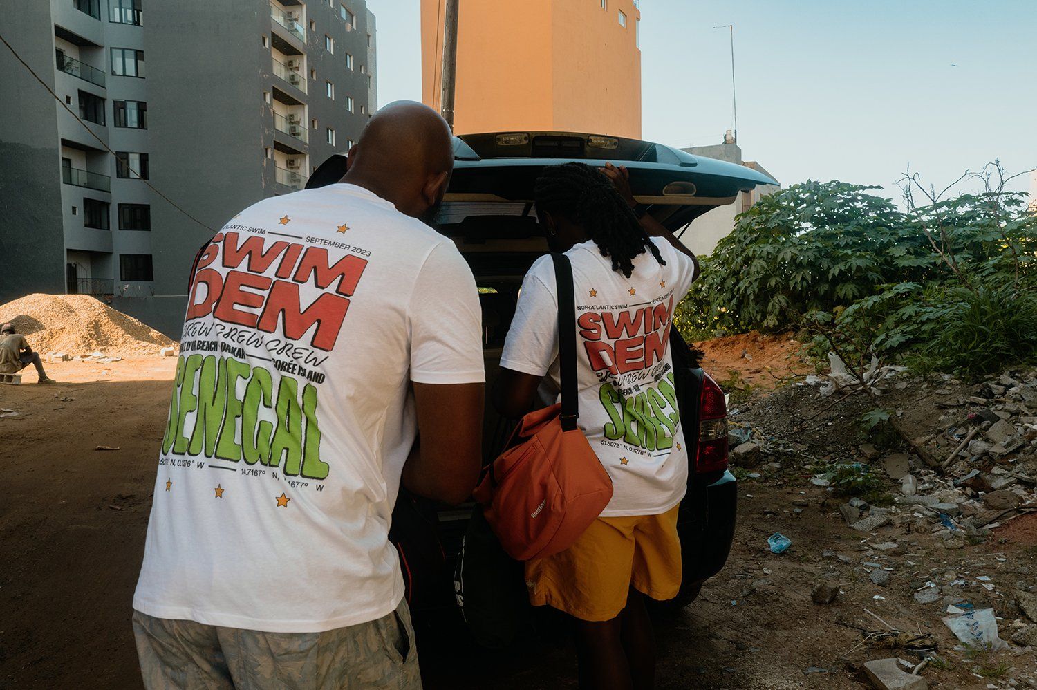 Two people unload the back of a van in white shirts that read \