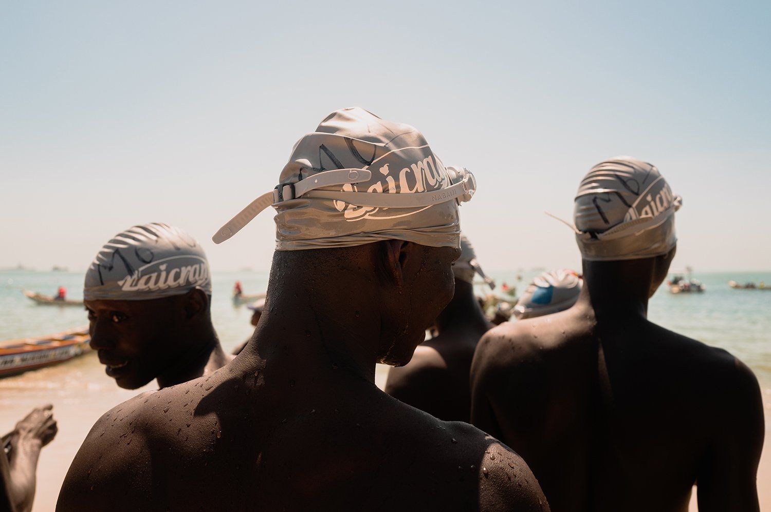 A group of swimmers in silver swim caps and silver goggles are seen from behind looking out at the water.