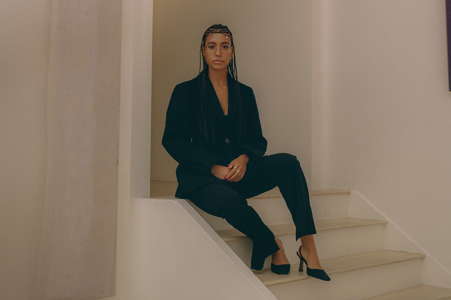 Hannah Traore is sitting on beige steps wearing a black suit and heels. Her hair is in thin braids adorned with jewelry at the front.