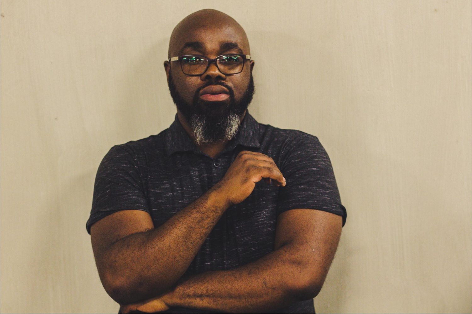 A Black man with a beard and glasses weares a dark colored polo shirt and poses for a portrait photo.
