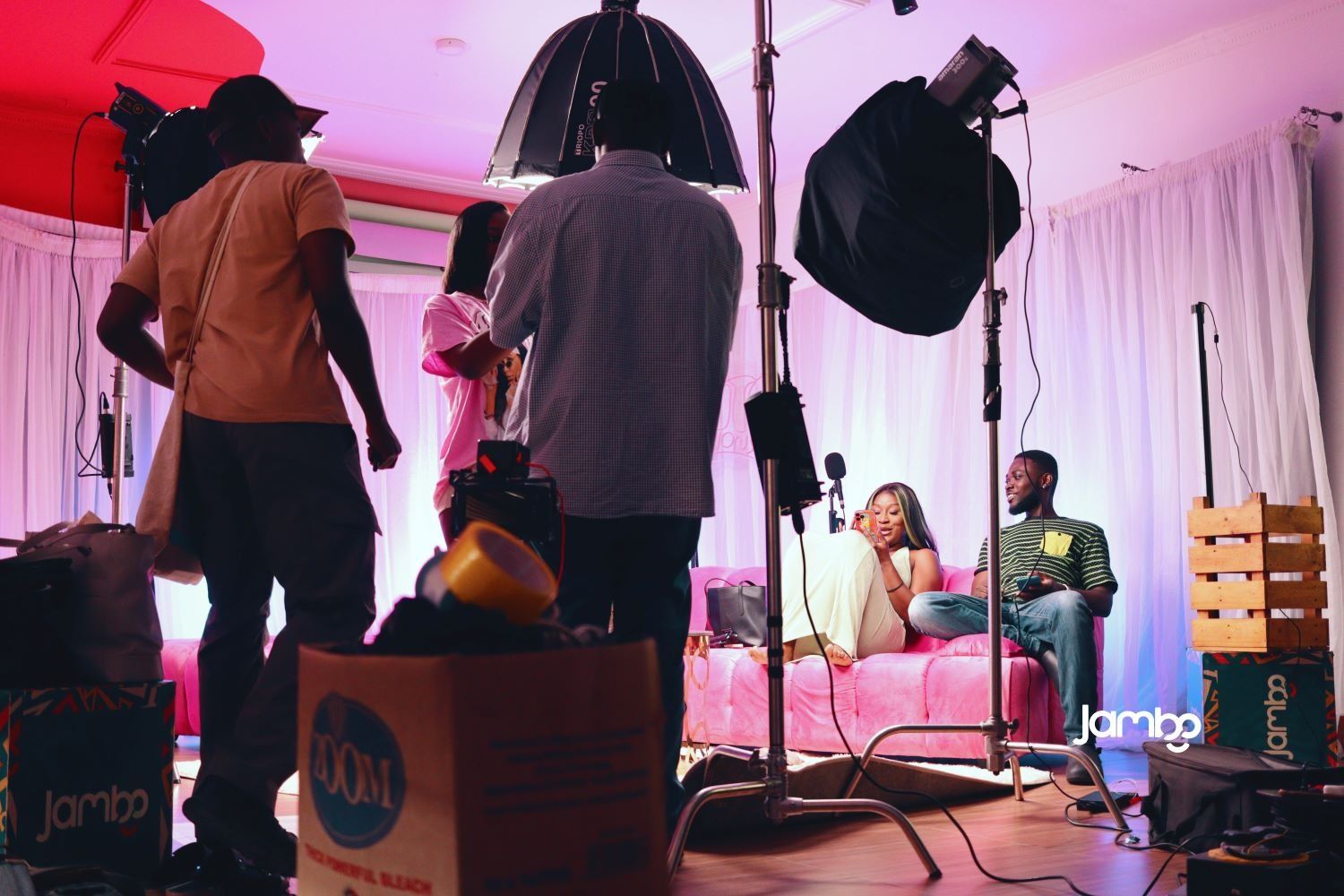 Behind-the-scenes of a video shoot, with crew members adjusting lighting and equipment while two people relax on a pink couch in the background.