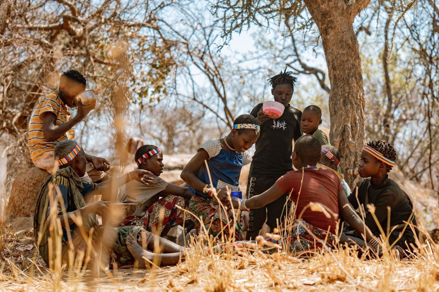 A group of eight young people sitting under trees, drinking from bowls. Some are wearing beaded head adornments over their short hair.