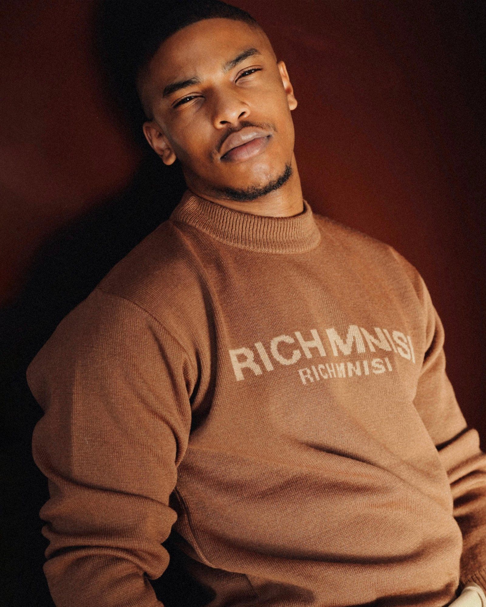 Actor Molaba in tan sweater against dark red background.