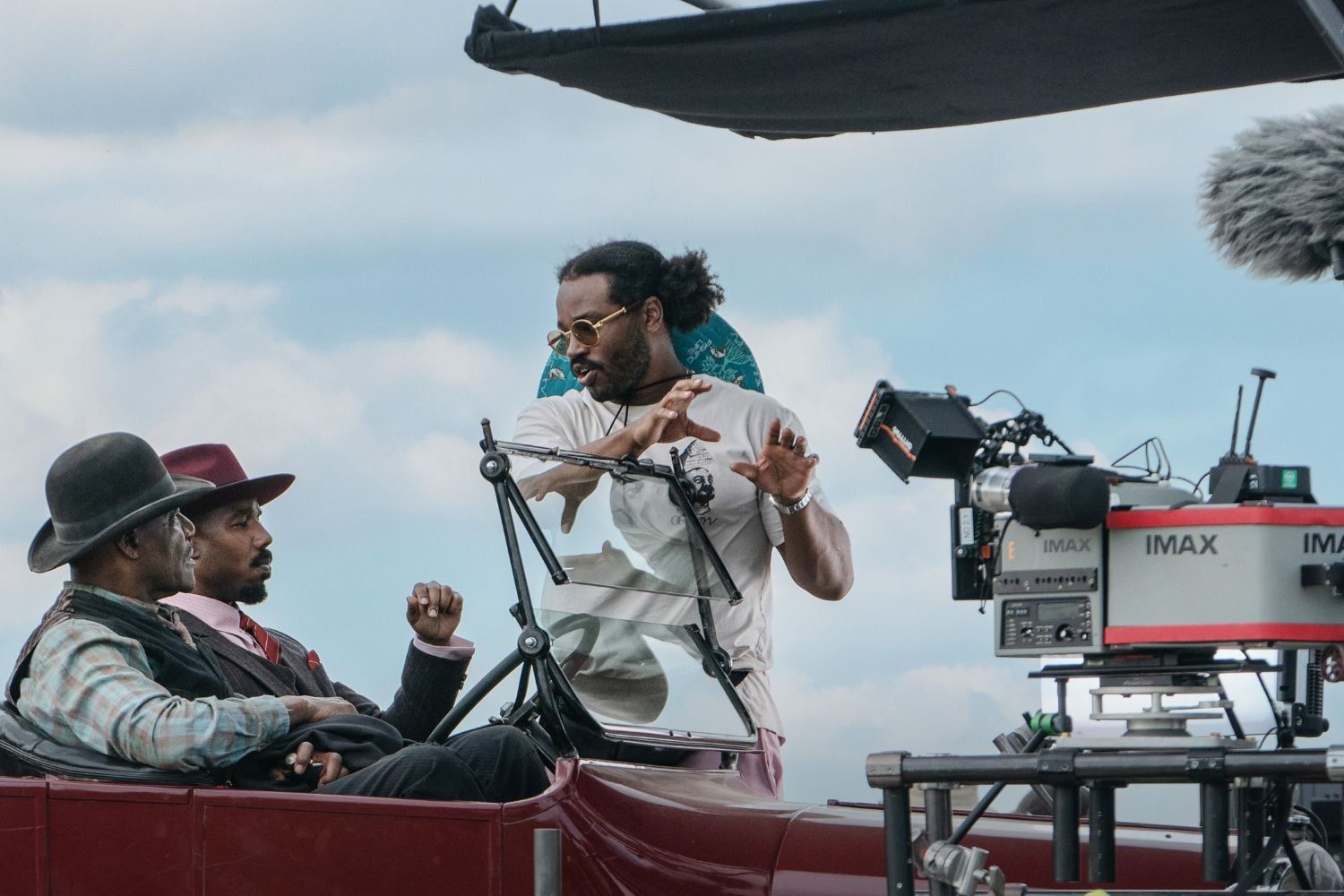 The actors Delroy Lindo and Michael B. Jordan are sitting inside a vintage car, looking at director Ryan Coogler as he gives them a direction on the set of the Sinners movie. There are Imax cameras in front of them.