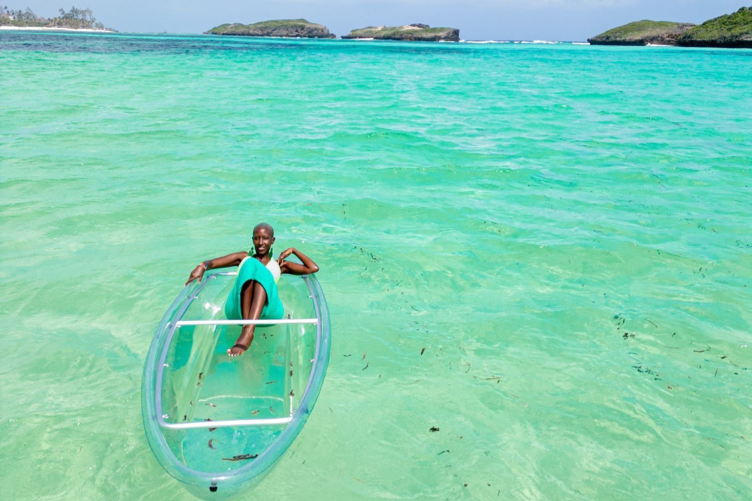 Gachuhi lays out in a transparent kayak on turquoise waters under a blue sky, surrounded by small islands.