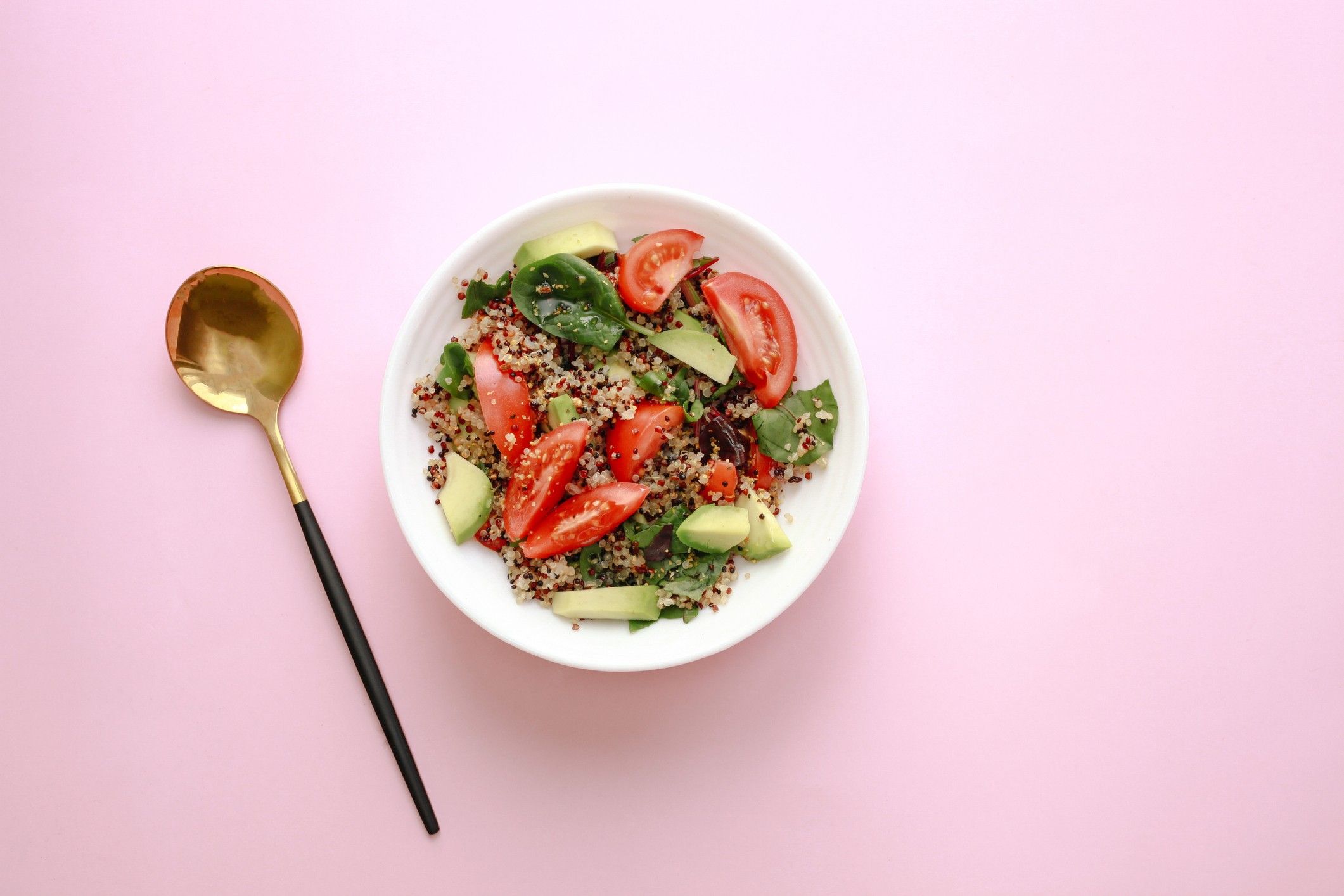Top-down quinoa salad with tomatoes, avocado, and leafy greens in a white bowl against a pastel pink background