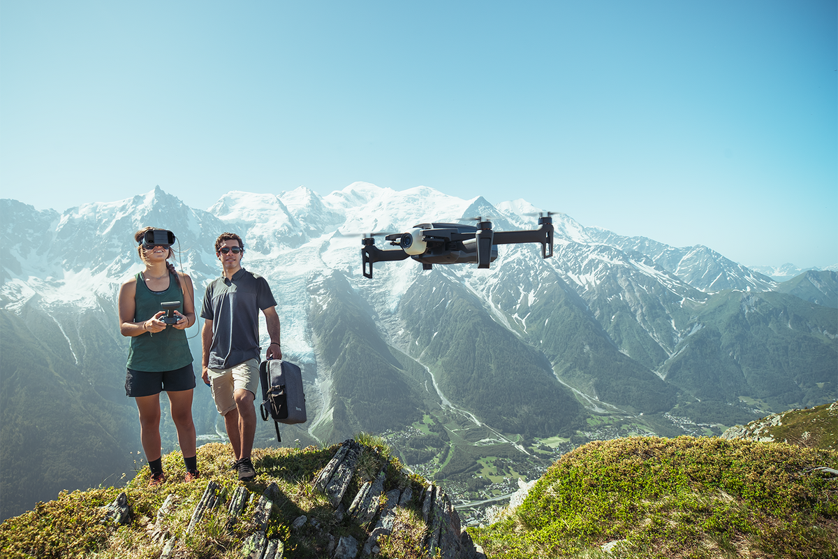 Two people flying a large drone in the foreground, with snow-capped mountains in the background