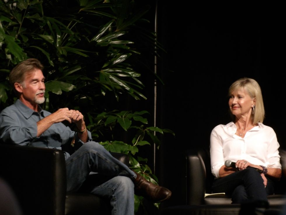 Amazon John Easterling and Olivia Newton-John sitting on black chairs with greenery in the background
