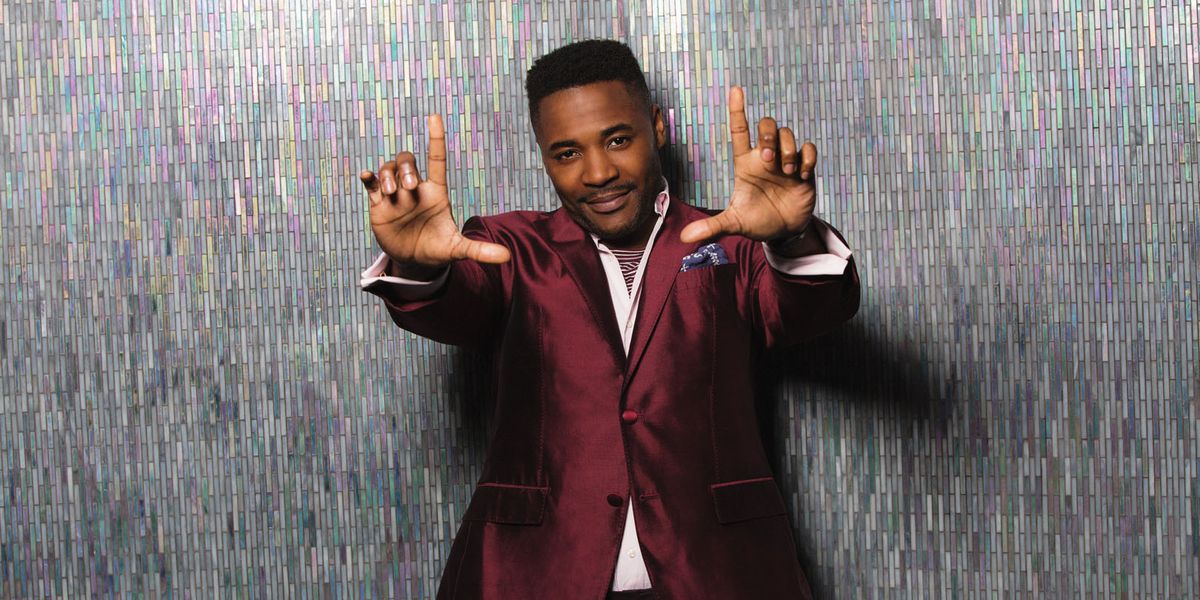 Duane Henry smiling in a burgundy colored suit.