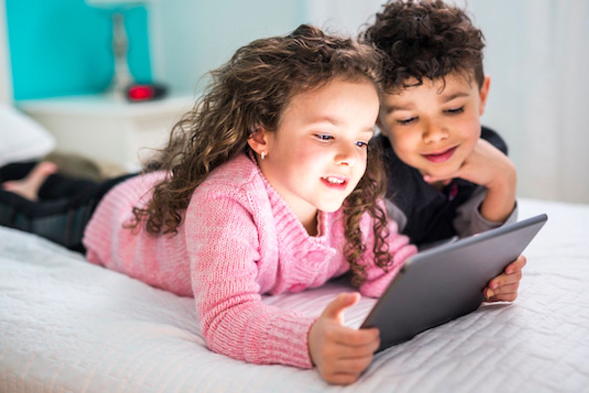 Two children lying on a bed looking at a tablet