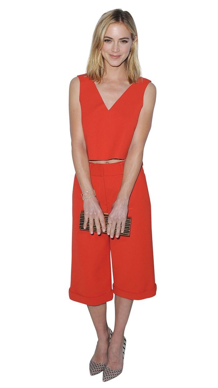Emily Wickersham in a cropped red shirt and capris holding a gold clutch