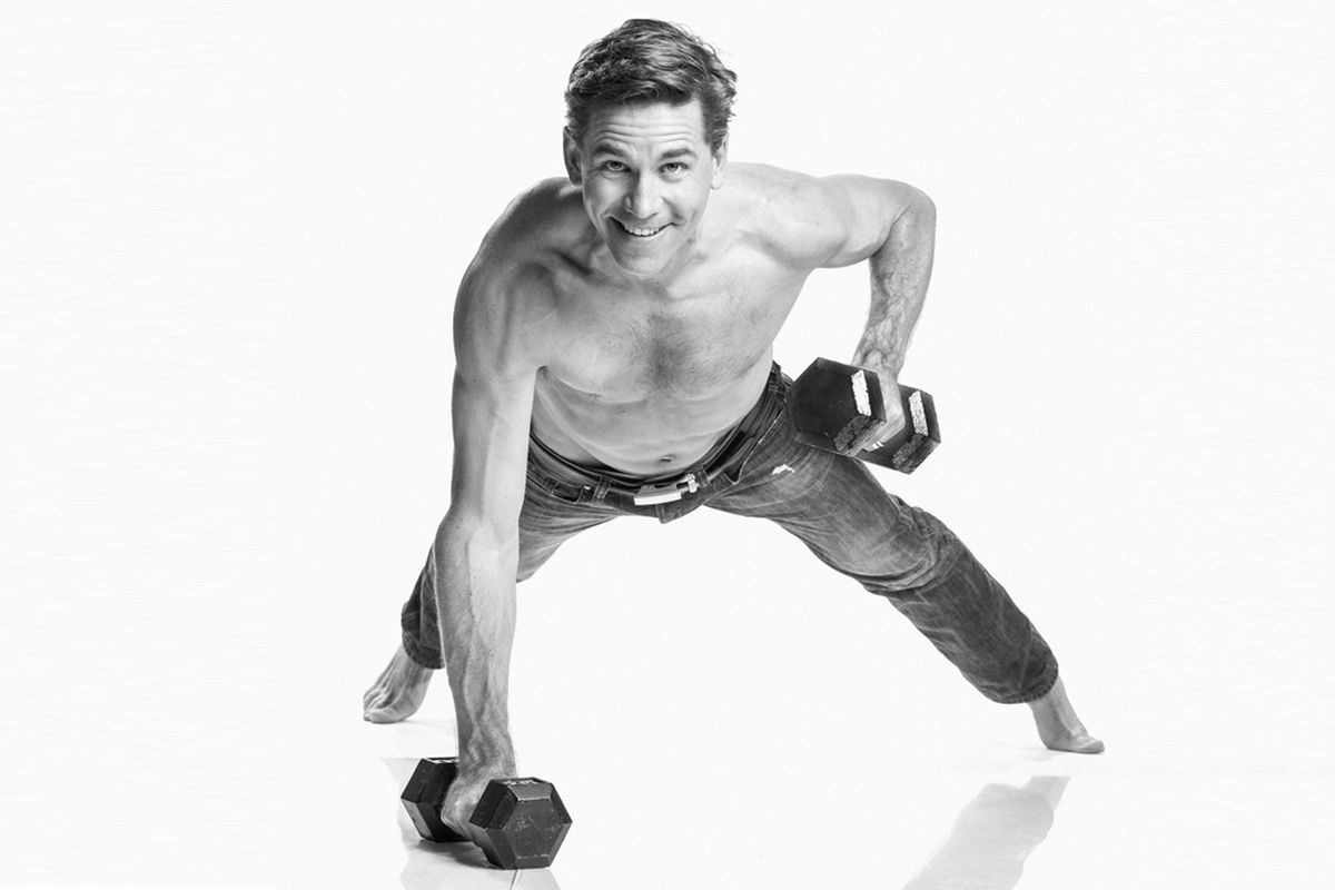 A black and white picture of a shirtless Brian Dietzen doing a one handed push-up with two weight in his hands