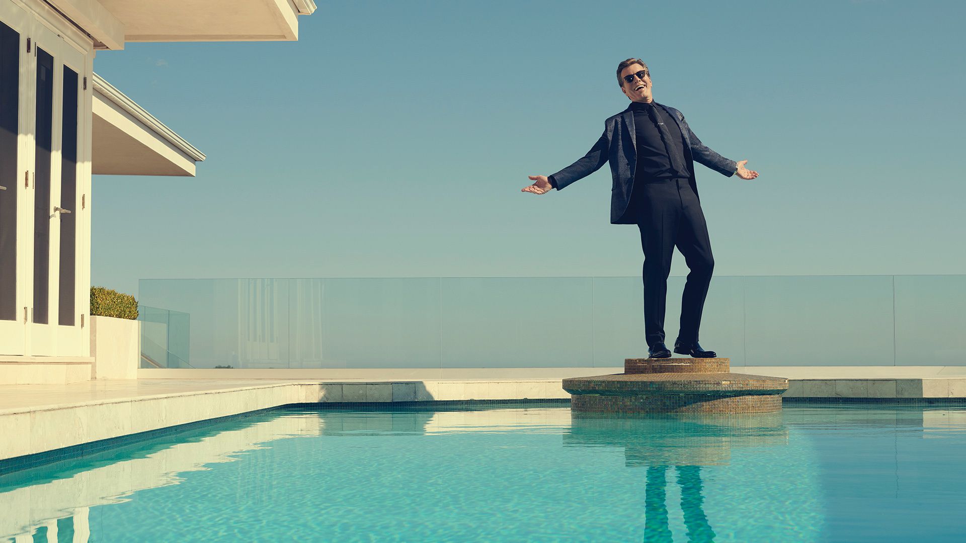Peter Bergman standing in a suit with his arms outstretched on a platform on an in-ground pool
