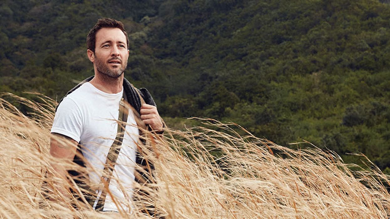 Alex O'Loughlin wears a white tee as he stands in a flowing field of wheat.