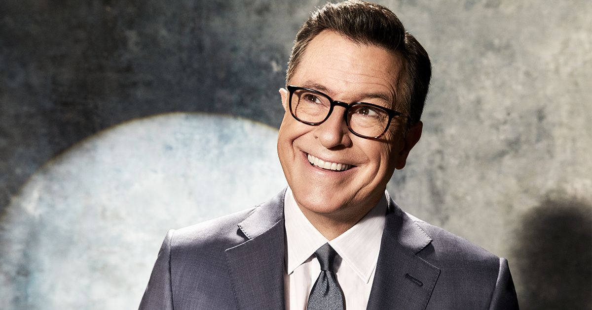 Close-up portrait of Stephen Colbert in a gray suit with a spotlight behind him.