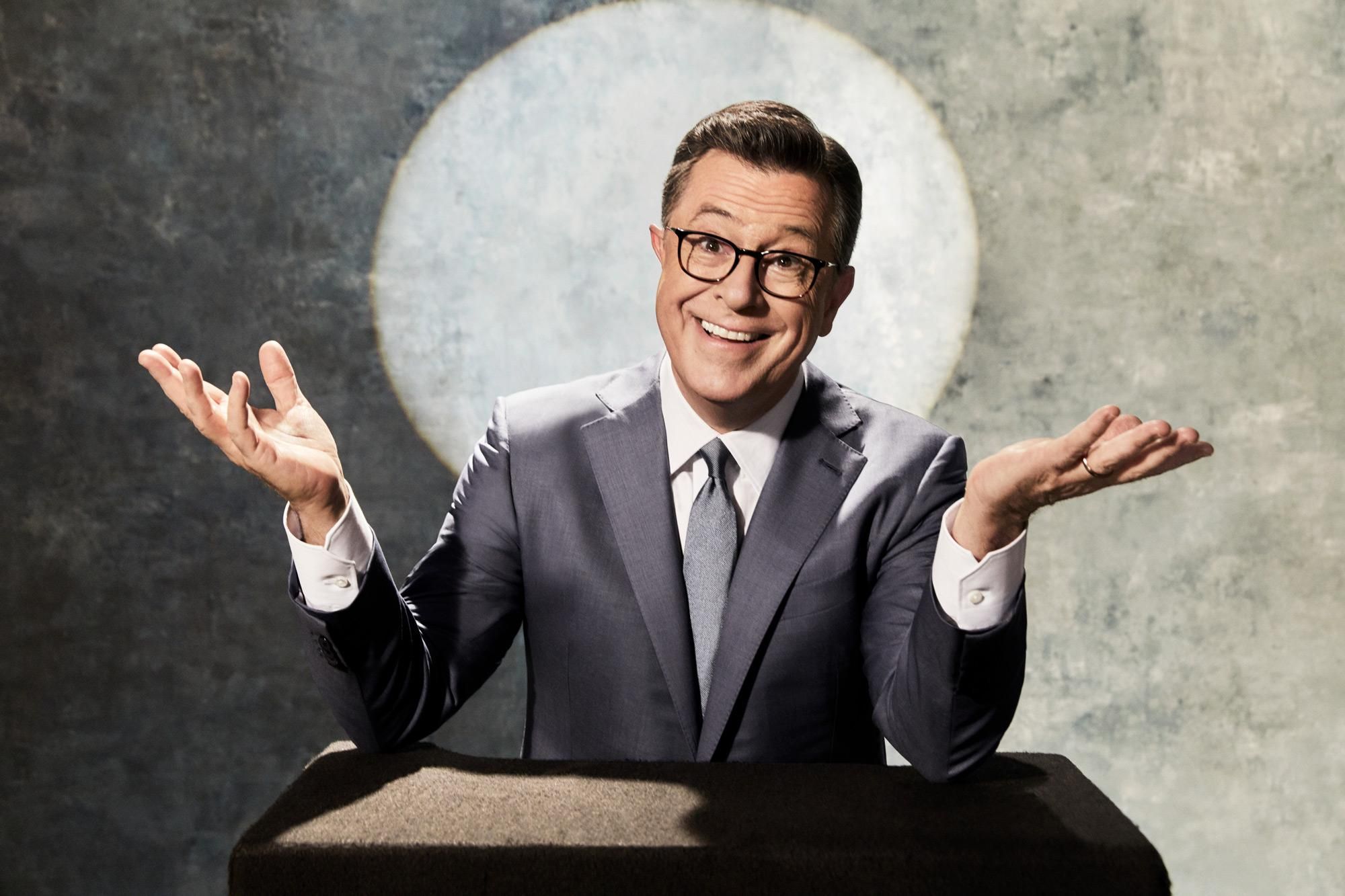 Stephen Colbert smiles broadly as he gestures from a carpeted podium.