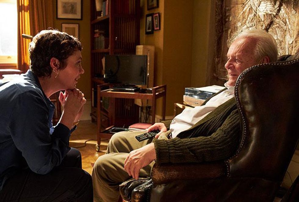 Olivia Colman and Anthony Hopkins are pictured in a still from a movie called "The Father." Colman (left) has short brown hair, and she's wearing a simple, moderately sized set of gold earrings. She wears a blue blouse. She's facing Hopkins, who plays her father, who is sitting on the right in a brown armchair. He wears a green cardigan with a white button up shirt and khakis while clutching a TV remote.