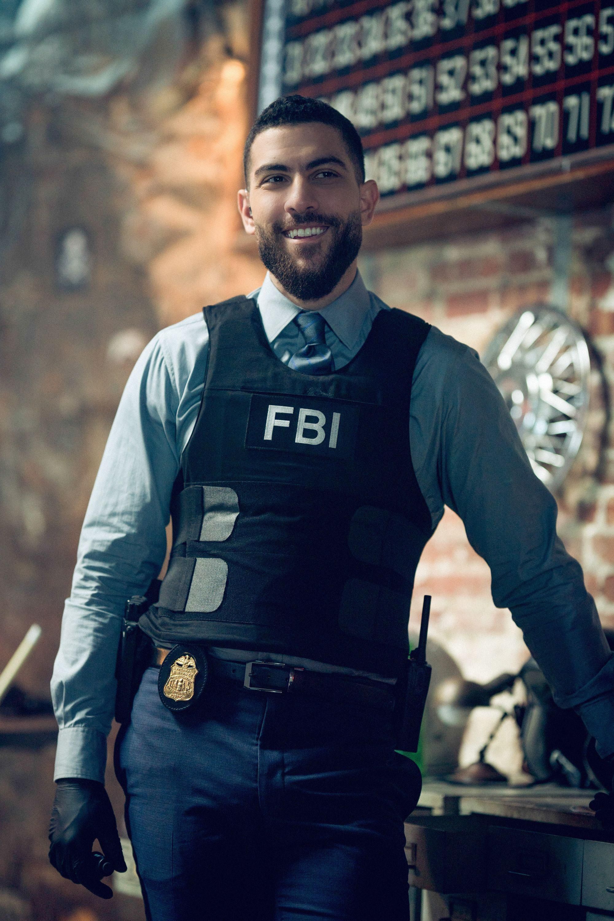Actor Zeeko Zaki leans against a counter and smiles broadly in his tactical FBI vest