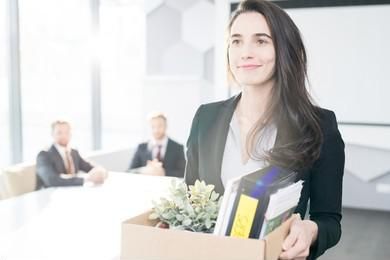 A woman holding a box filled with plants, binders and papers. She is dressed in a blazer and blouse, and has a small smile on her face.