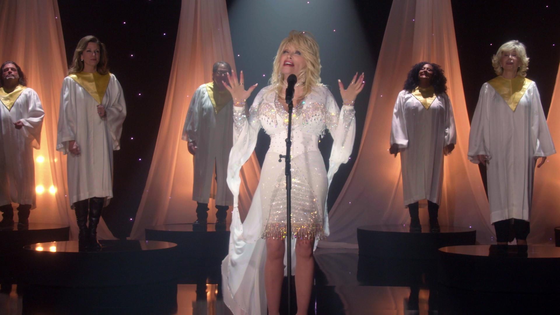 Dolly Parton stands onstage at a microphone wearing a white dress with gold trim while backed by choir singers in white robes