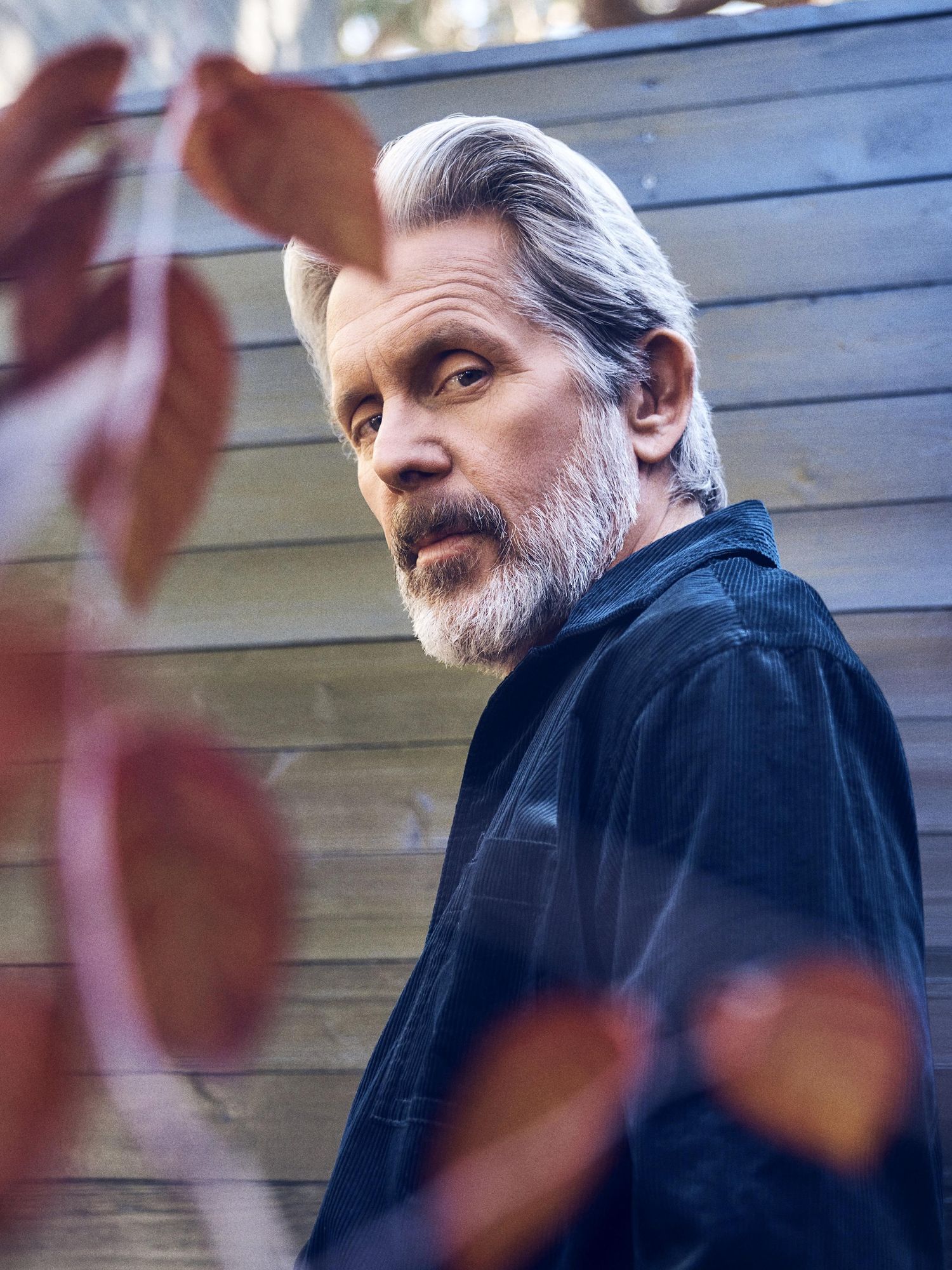 Actor Gary Cole from NCIS leans against a wooden fence wearing a dark blue corduroy jacket.