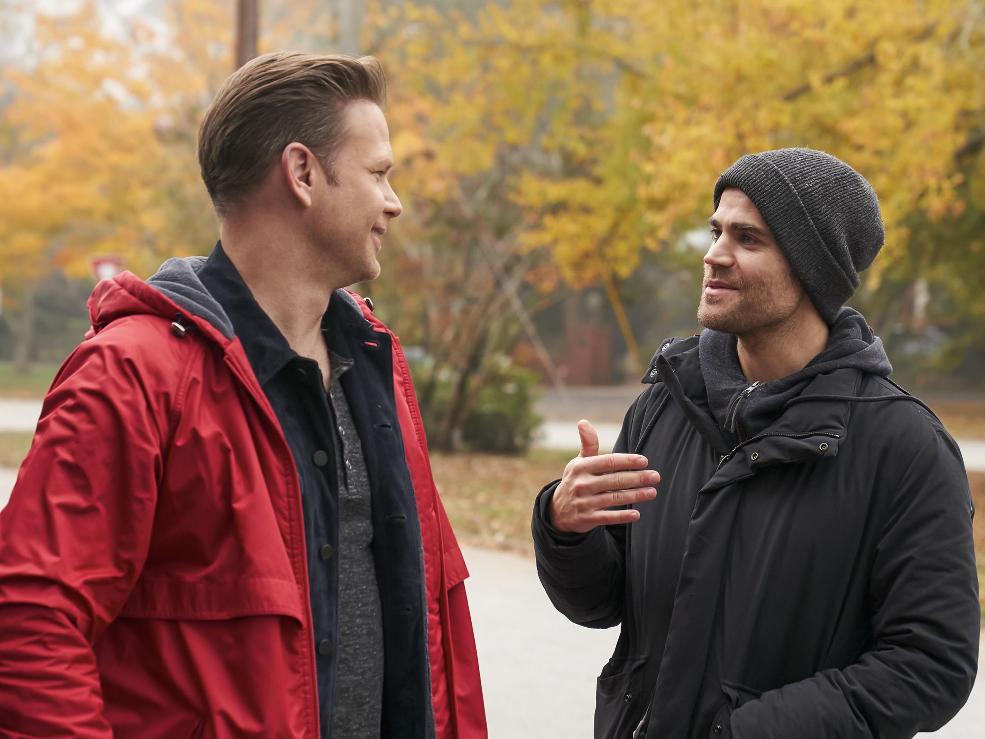 Paul Wesley and Matthew Davis smile and chat near a line of trees covered in fall foliage on the set of Legacies