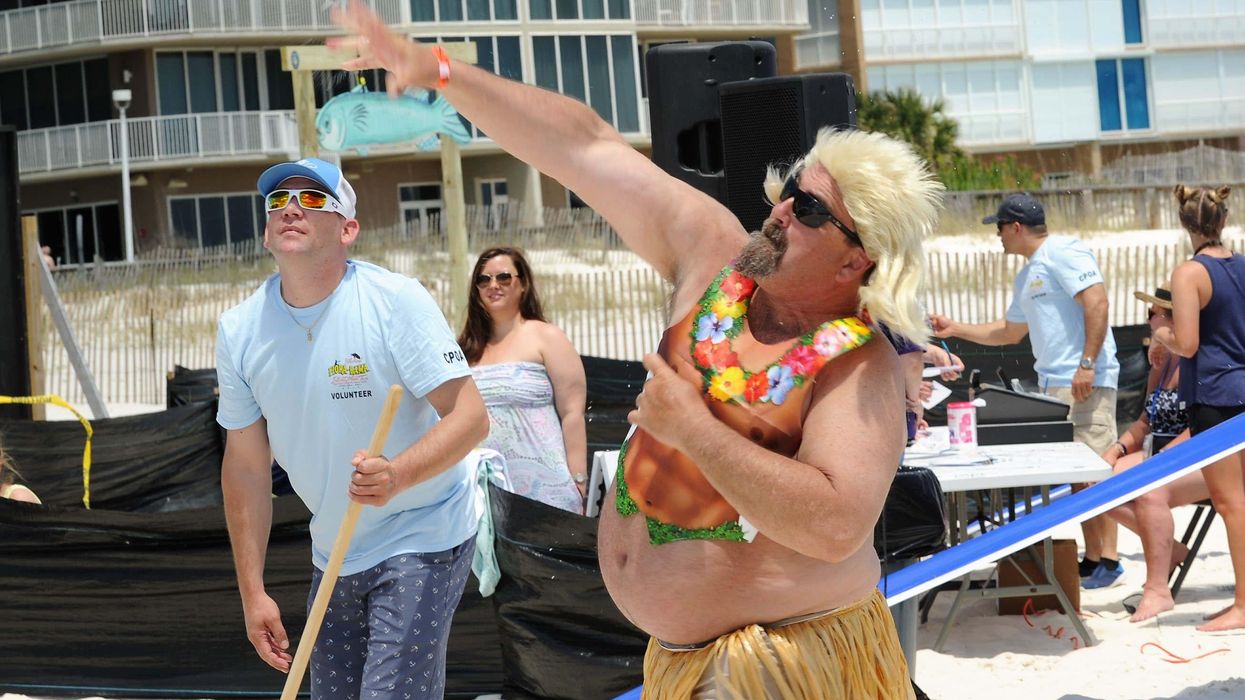 Man in a blond wig and grass skirt throws a mullet fish with people looking on