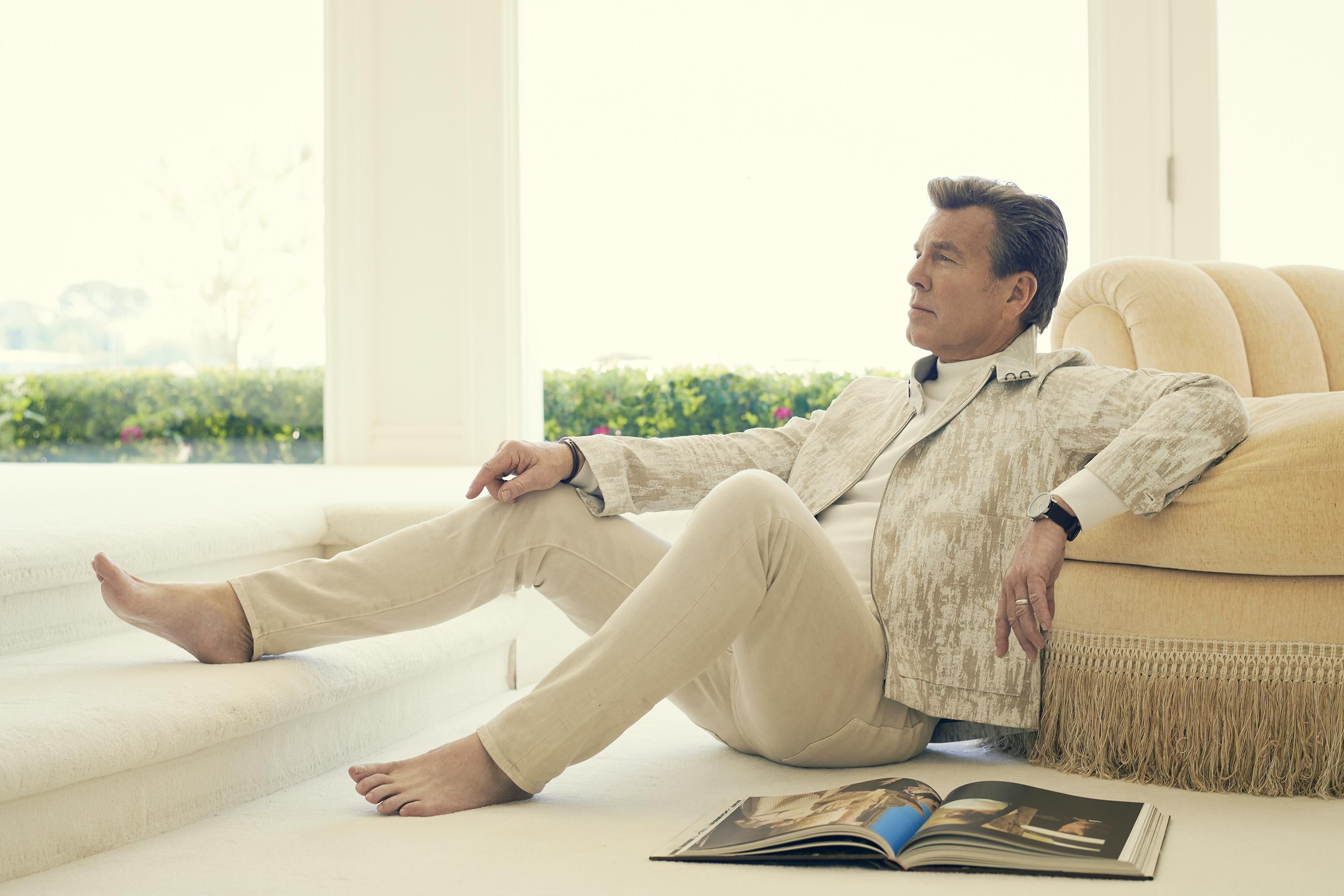 Peter Bergman wears a beige jacket and pants while sitting on a cream carpet next to an open art book. He leans his left arm against a chair and looks into the distance
