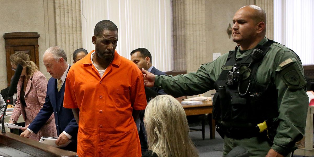 A Black man, R. Kelly, stands in a court room, wearing an orange jumpsuit with his hands handcuffed behind his back, accompanied by a police officer in a green uniform, bulletproof vest and gun.