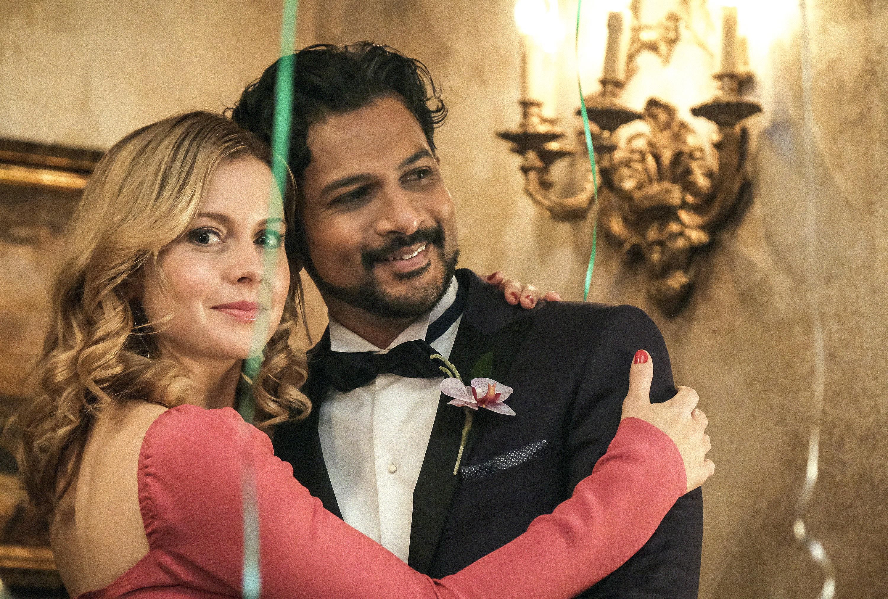 Rose McIver as Samantha wears a salmon pink dress and puts her arms around a tuxedo clad Utkarsh Ambudkar as Jay in Ghosts