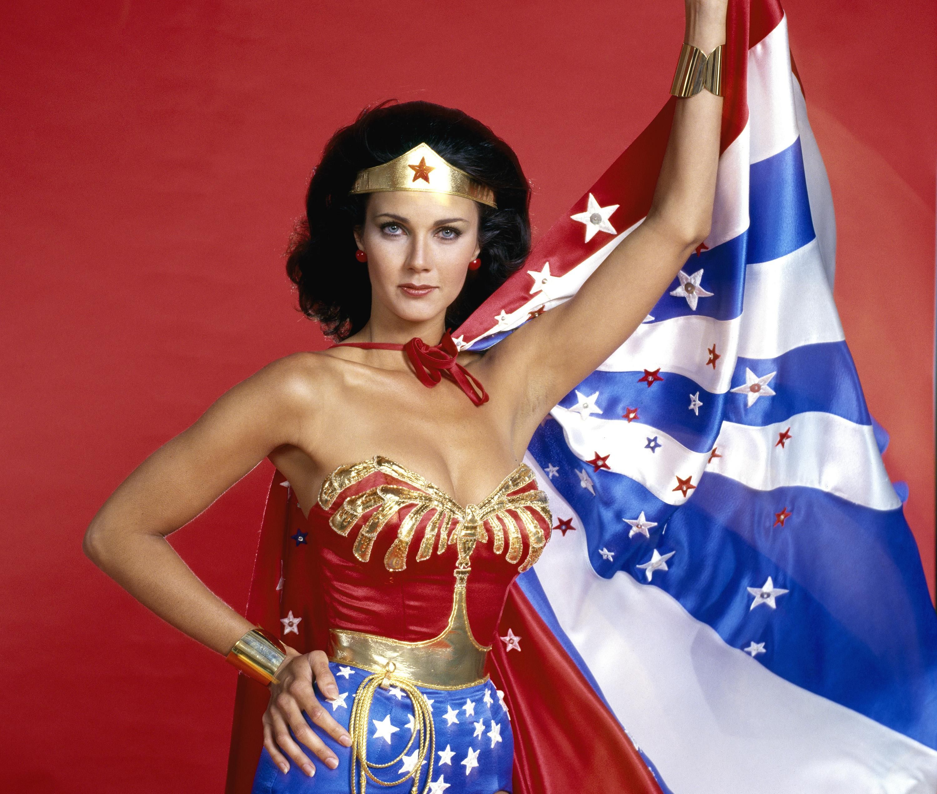 Actress Lynda Carter dressed as Wonder Woman in a star-spangled outfit with a golden crown and a blue and white cape that she holds up with her left hand