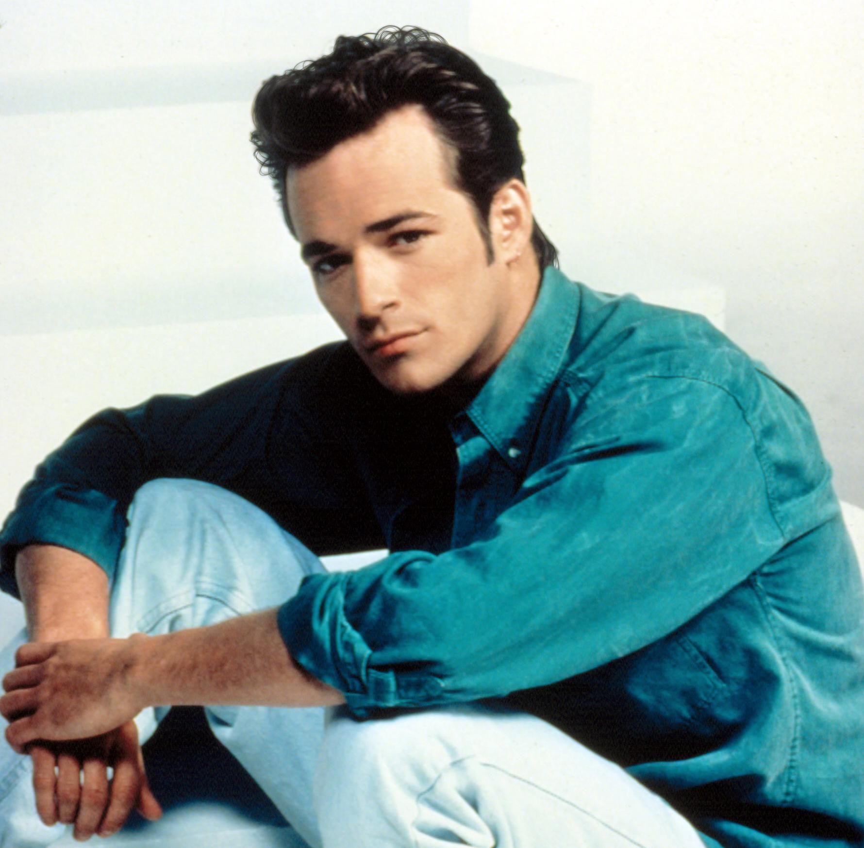 Actor Luke Perry in light blue jeans and a teal collared shirt sitting on some steps giving a smoldering look.