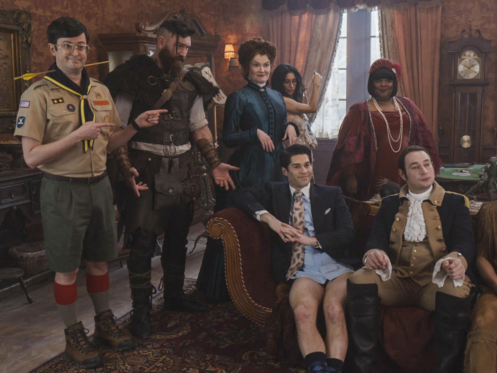 The cast of Ghosts sitting on and standing around a couch