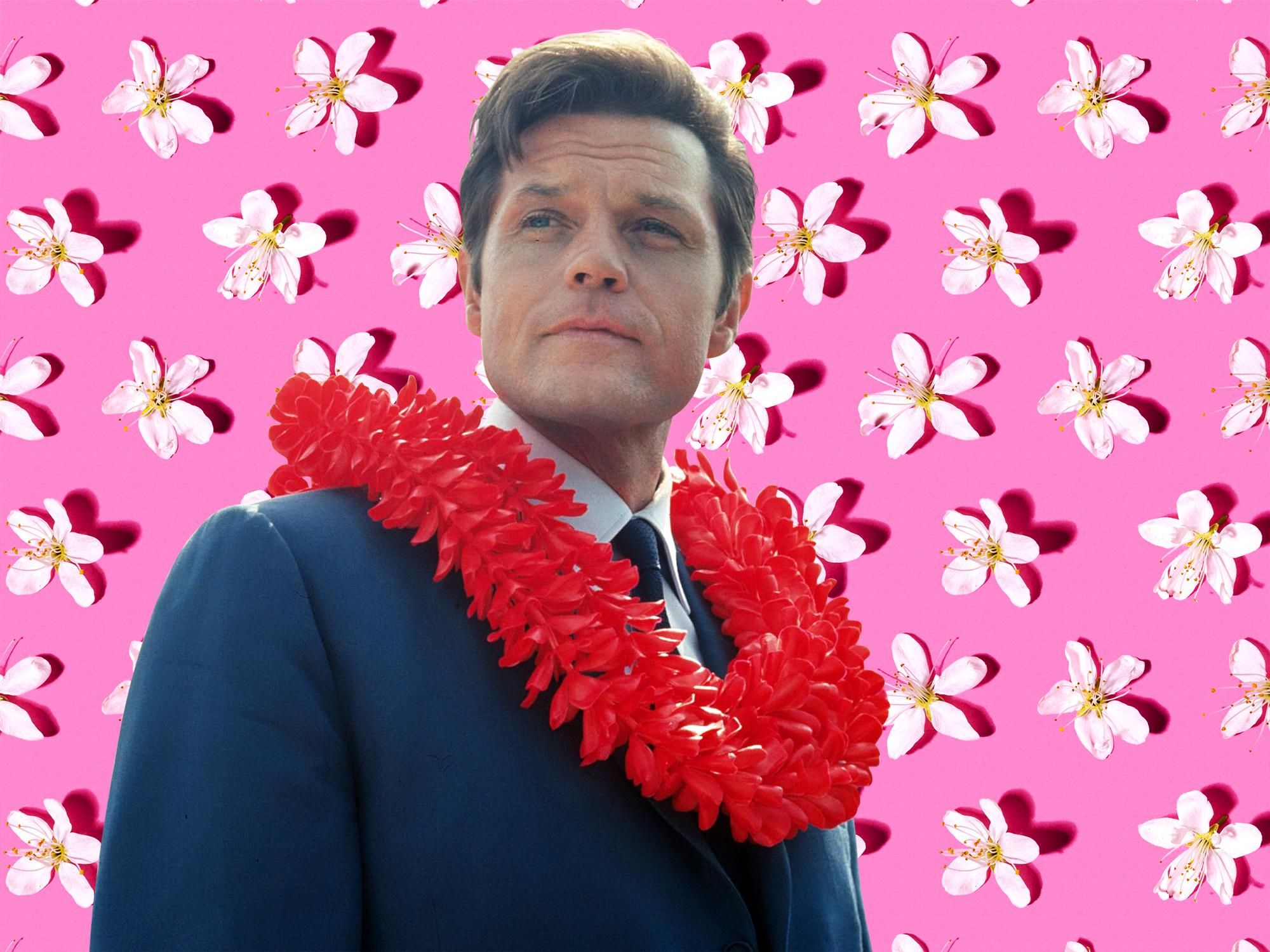 Jack Lord of Hawaii Five-O poses against a pink floral backdrop while wearing a navy suit with a bright red flower lei around his neck.