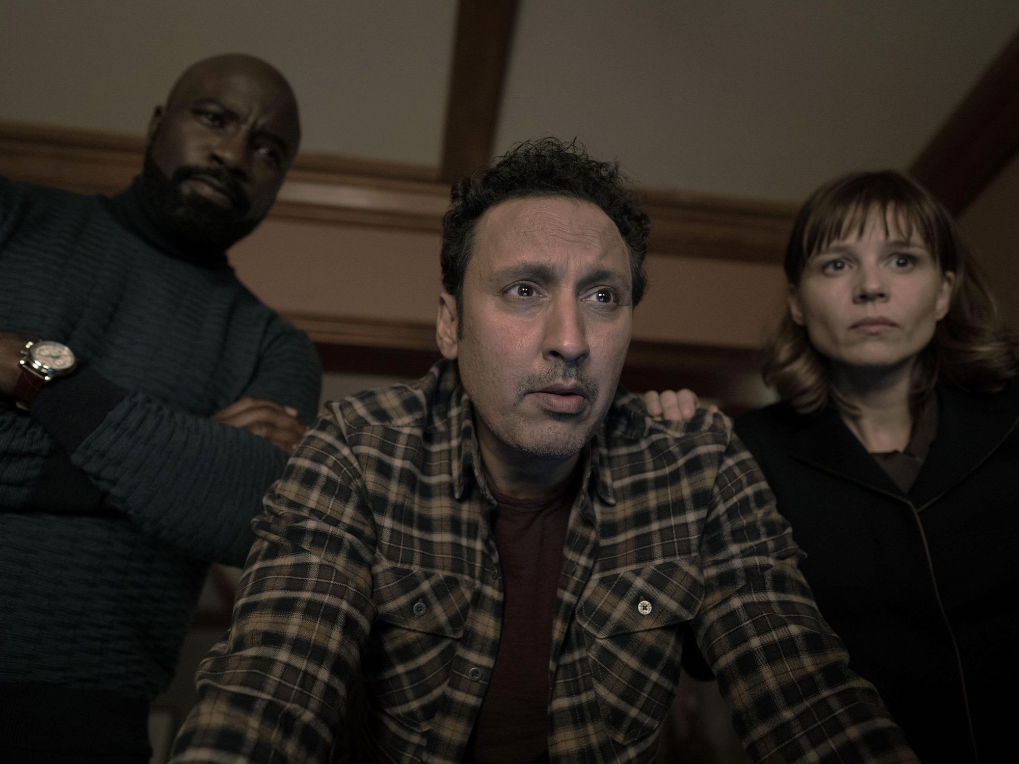 Aasif Mandvi wears a brown plaid shirt and stands between Mike Colter in a blue sweater and Katja Herbers in black while all three stare ahead fearfully