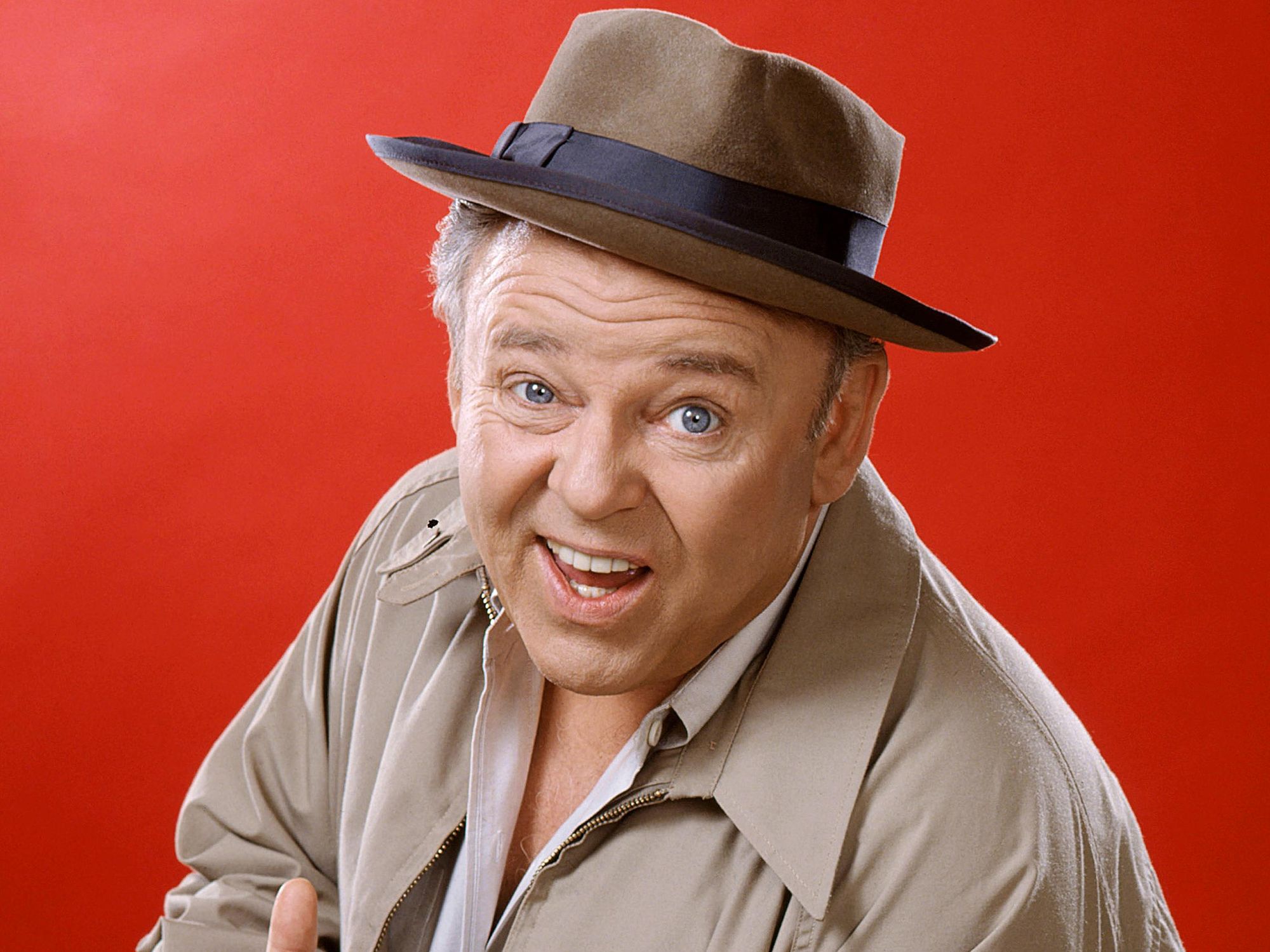 Carroll O'Connor of All in the Family, wearing a brown hat with a dark blue band and a beige jacket, against a red background.