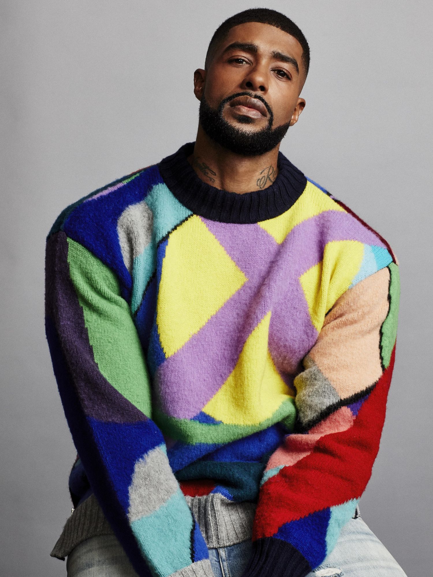 Skyh Alvester Black seated and shown from the waist up wearing a bright multicolored pullover sweater.