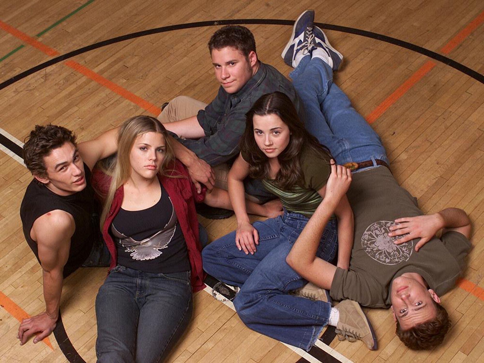 James Franco, Busy Phillips, Seth Rogan, Linda Cardellini, and Jason Segal sit on a gym floor staring up the camera.