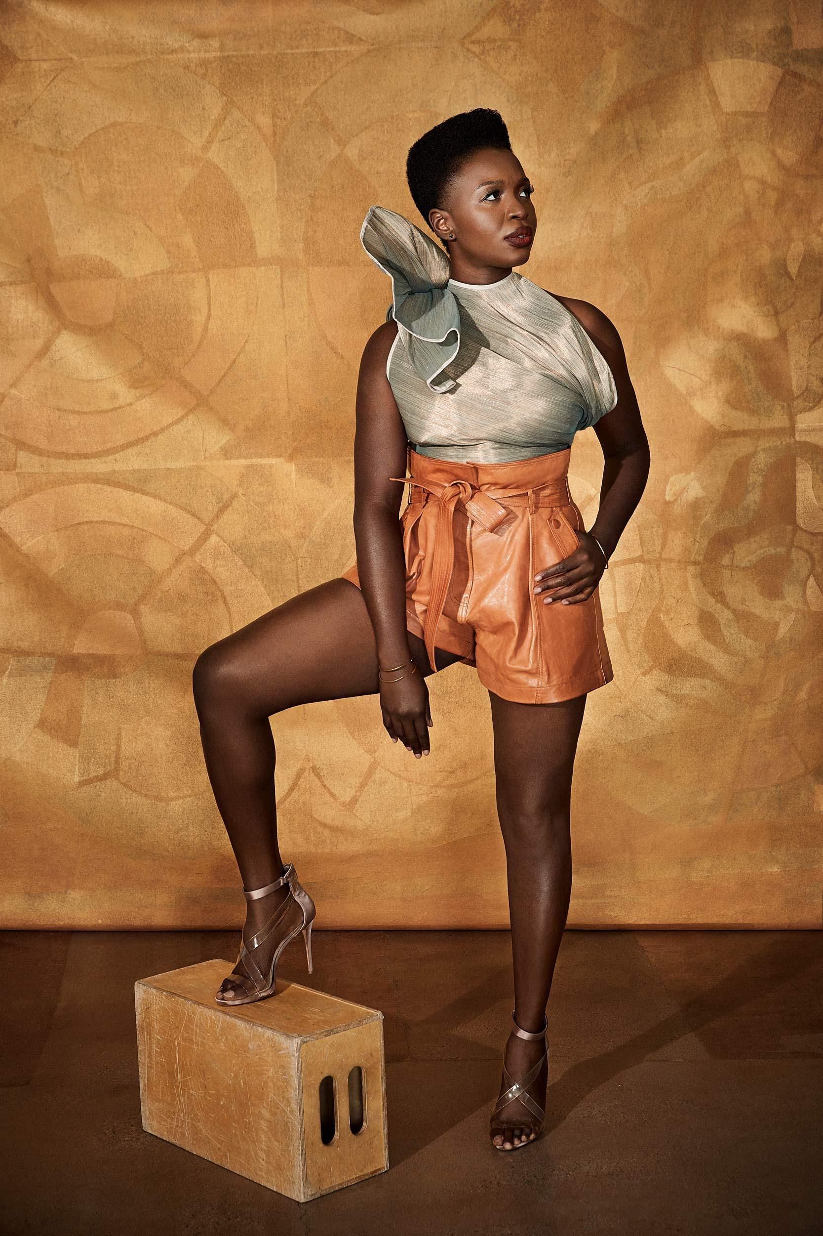 Folake Olowofoyeku poses with one foot on a wooden box while wearing tan leather shorts and a sleeveless gray blouse with a large ruffle.